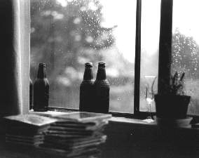 Black and white. Root beer bottles on the windowsill.  Rain-spotted window.  Some CDs piled up.
